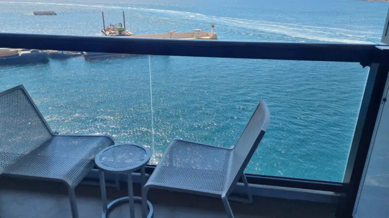 Balcony of a Virgin Voyages cruise ship cabin with two metal chairs and a table, overlooking the ocean and distant hills.