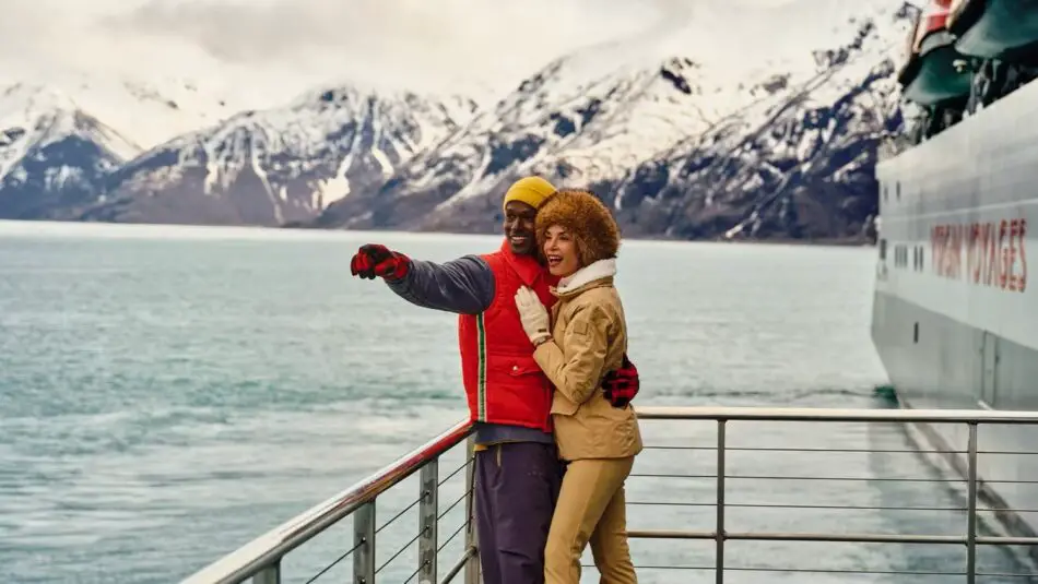Smiling couple on the deck of a Virgin Voyages cruise ship with snow-capped mountains in the background