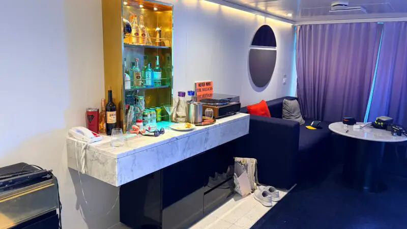 Interior of a Virgin Voyages cruise ship cabin featuring a marble-topped bar area with assorted bottles and glasses, a seating area with a dark sofa and round table, and curtains covering a window or balcony door.