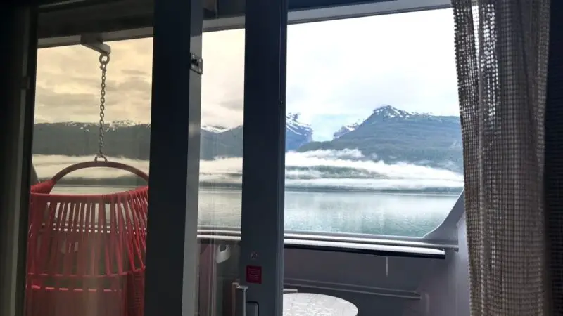 Virgin Voyages cruise ship cabin balcony with a hanging red chair, small white table, and scenic view of mountains and water through sliding glass doors.