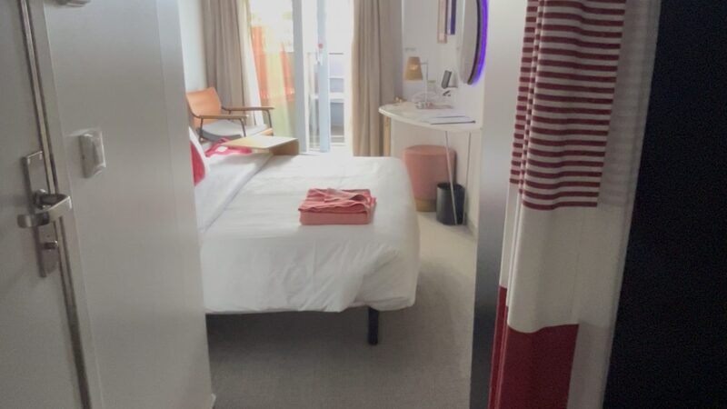 Virgin Voyages cruise ship cabin with a bed, desk, chair, and door leading to a balcony.
