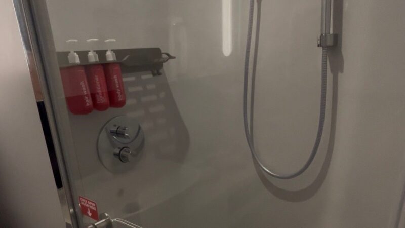 A Virgin Voyages cruise ship cabin shower with a glass door, chrome handle, and three mounted red toiletry bottles.