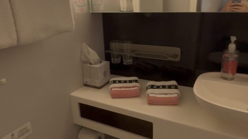 Bathroom in a Virgin Voyages cruise ship cabin, featuring a toilet, sink, mirror, and towels.