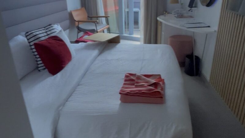 Virgin Voyages cruise ship cabin with a bed, pillows, and a folded towel. There is a desk with a mirror and a chair by the balcony door.