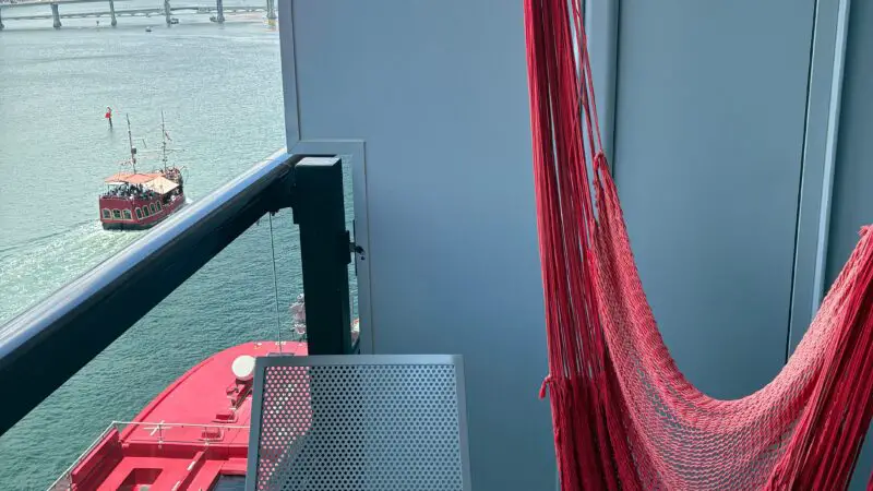 Virgin Voyages cruise ship balcony with a red hammock and gray chair overlooking the ocean and city skyline.