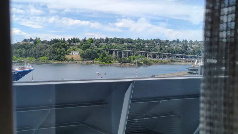 View from a Virgin Voyages cruise ship cabin balcony, showing a river, green hills, and a bridge under a blue sky.