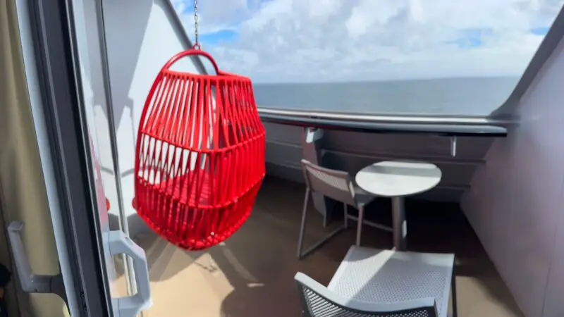 Cruise ship stateroom balcony with a red hanging chair, small white table, two chairs, and ocean view.