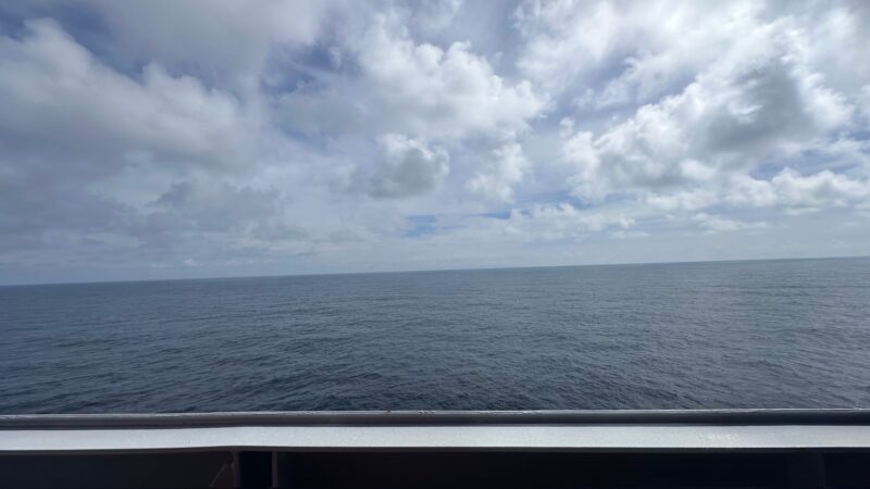 View from a Virgin Voyages cruise ship balcony, overlooking a vast ocean under a partly cloudy sky.
