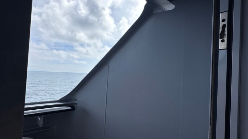 Balcony view from a Virgin Voyages cruise ship cabin, showing a chair and table with a partial ocean and sky view.