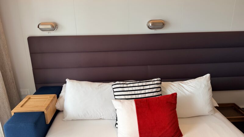 Virgin Voyages cruise ship cabin with a modern headboard, white bedding, striped and red accent pillows, and integrated bedside lighting.