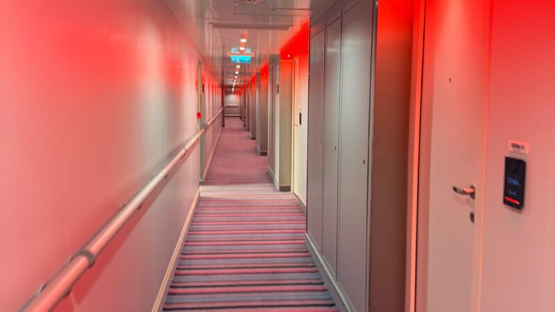 A cruise ship corridor on a Virgin Voyages ship with striped carpet, white walls, and red lighting overhead.