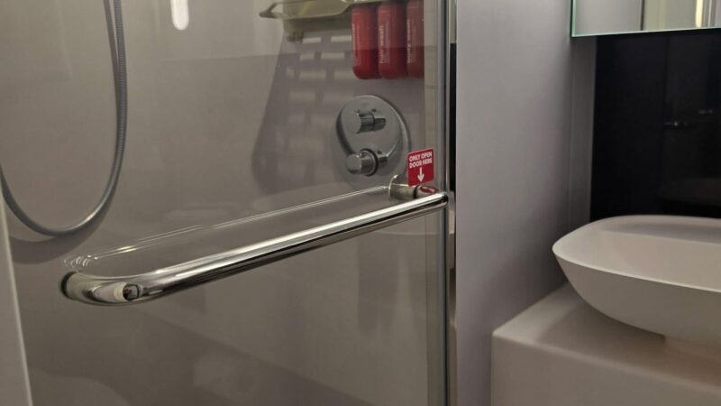 Cruise ship cabin bathroom featuring a shower with a glass door, wall-mounted toiletries, and a basin sink next to shelving.