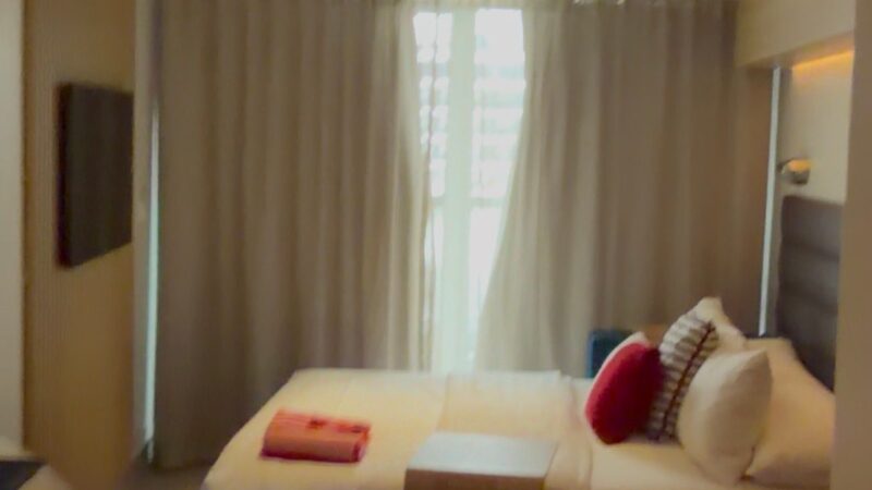 Virgin Voyages cruise ship cabin with a bed, pillows, and a blanket, featuring a balcony with curtains.