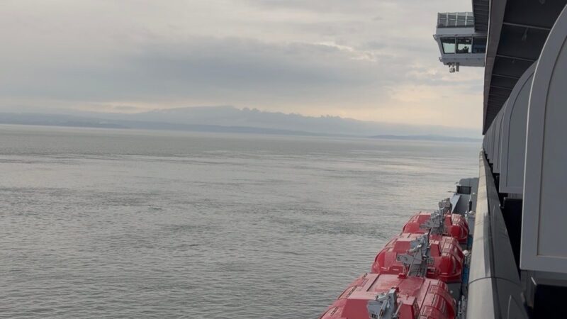 View from a Virgin Voyages cruise ship balcony, showing the ocean and several red lifeboats along the side of the ship. Overcast sky in the background.