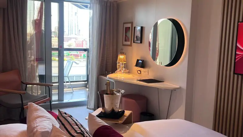 Virgin Voyages cruise ship cabin with a bed, pillows, a wall mirror, a desk with a lamp, and a balcony with outdoor seating.