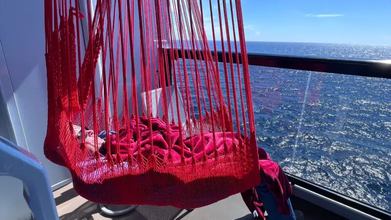 Cruise ship balcony with a red hammock-style chair overlooking the ocean under a clear blue sky.