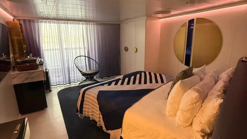 Interior view of a Virgin Voyages cruise ship cabin featuring a bed with decorative pillows, a modern chair, a round side table, and soft ambient lighting.