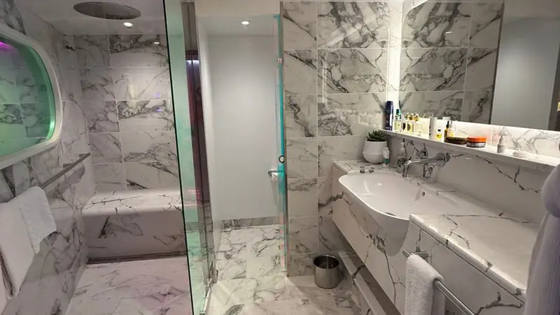 Luxurious Virgin Voyages cruise ship stateroom bathroom with marble tiles, dual sinks, and a glass-enclosed shower.