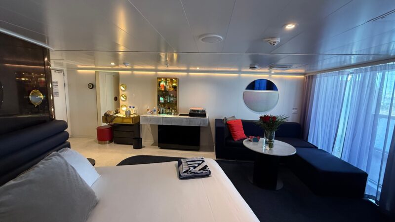 A Virgin Voyages cruise ship cabin with a bed, seating area, round table with flowers, and a balcony with sheer curtains.
