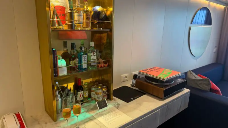 A Virgin Voyages cruise ship cabin featuring a well-stocked minibar on a marble counter, with two glasses and a record player beside it. A round window and sofa are in the background.