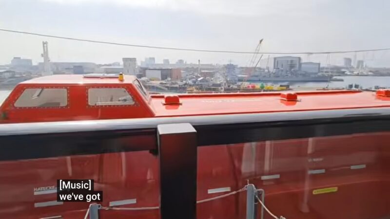 Balcony view from a Virgin Voyages cruise ship cabin, featuring a red lifeboat and a harbor cityscape.