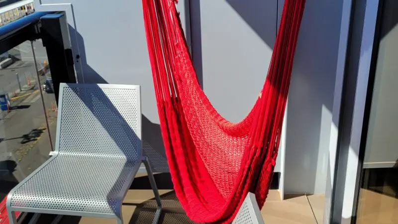 Cruise ship stateroom balcony with a red hammock, two white perforated chairs, and a small round table.