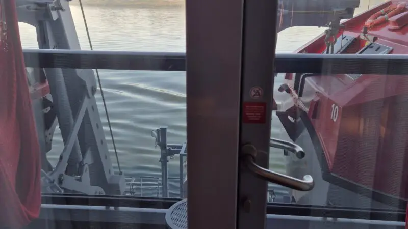 View from a Virgin Voyages cruise ship cabin balcony, showing a life raft and calm sea with distant hills.