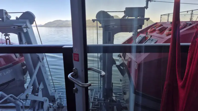 View from a Virgin Voyages cruise ship cabin balcony, showing a lifeboat and calm sea with hills in the background.