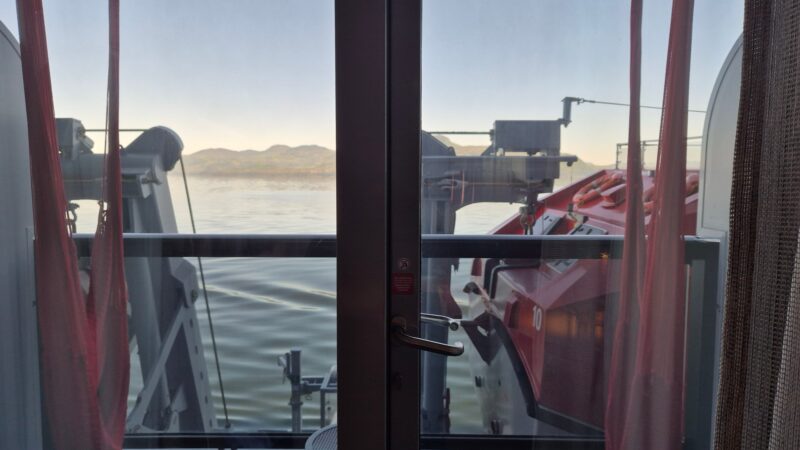 View from a Virgin Voyages cruise ship cabin balcony, showing lifeboats, calm water, and mountains in the distance through glass doors.
