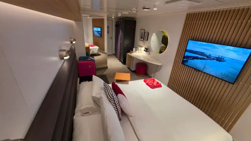 A modern Virgin Voyages cruise ship cabin featuring a neatly made bed, a wall-mounted TV, a desk with a round mirror, and stylish decor accents.