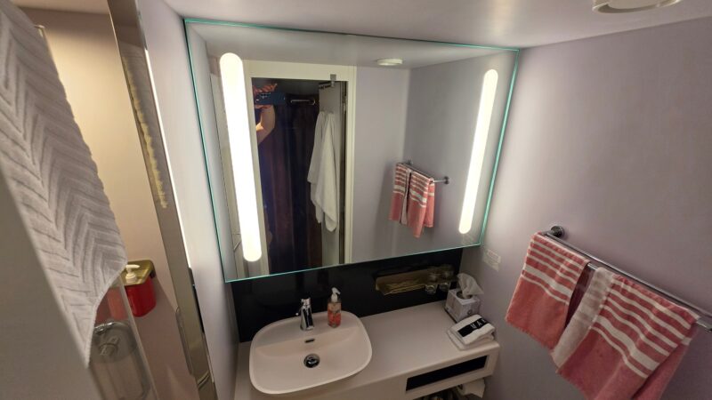 Bathroom of a Virgin Voyages cruise ship cabin with a sink, mirror, and striped towels.