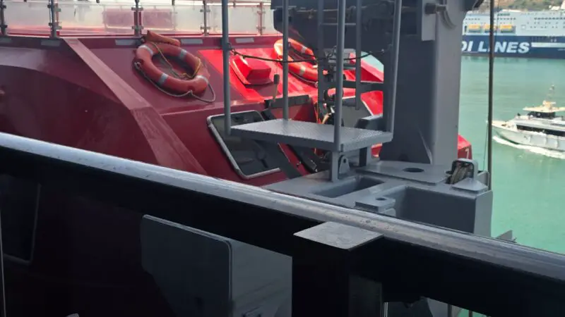 Balcony view from a Virgin Voyages cruise ship cabin, showing a red lifeboat, railings, and part of the ship's structure against a backdrop of the sea and distant boats.