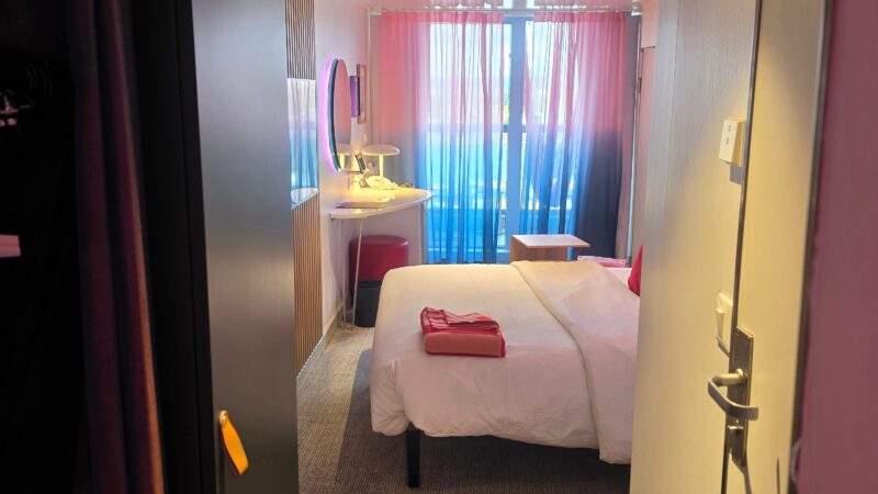 Virgin Voyages cruise ship cabin interior with a bed, pink and blue curtains, and a desk with a circular mirror.