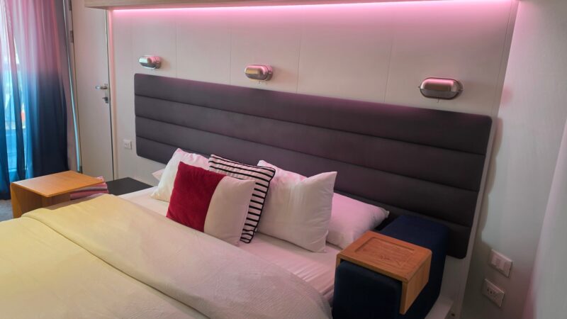 Virgin Voyages cruise cabin with a modern bed, decorative pillows, wood accents, and ambient pink lighting.