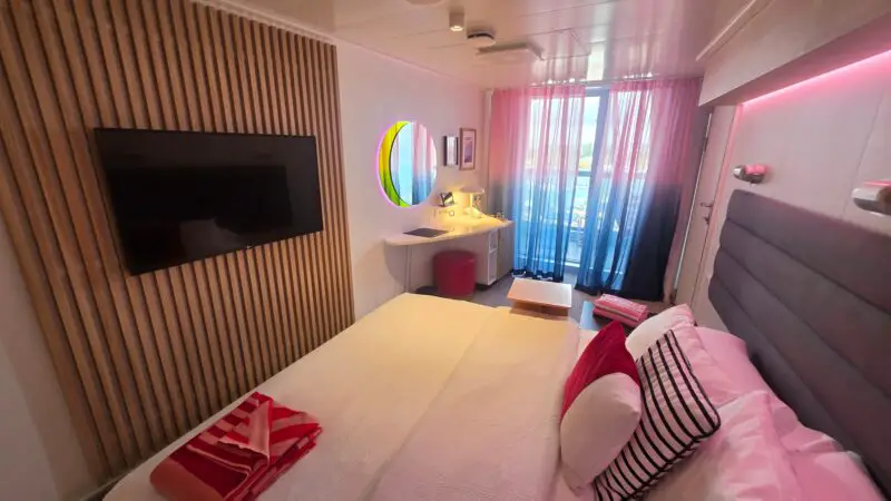 Virgin Voyages cruise ship cabin with a double bed, colorful pillows, wall-mounted TV, modern vanity, and glass doors opening to a balcony with sheer curtains.