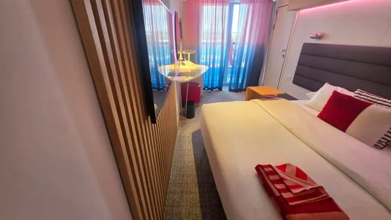 Virgin Voyages cruise ship cabin with a plush bed, vibrant pillows, a TV on a textured wall, and a balcony with sheer pink and blue curtains.
