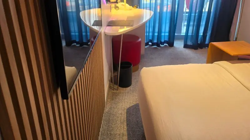 A Virgin Voyages cruise ship cabin featuring a bed, desk with a lamp, and pink and blue sheer curtains covering a balcony door.