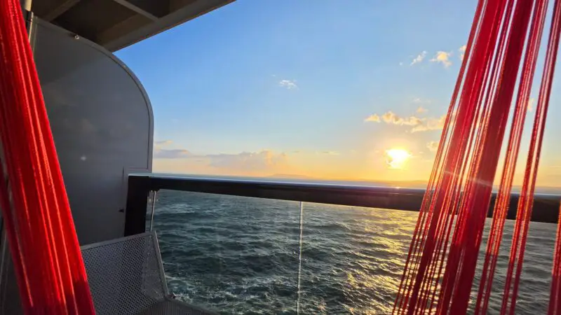 Virgin Voyages cruise ship balcony with a white chair and red hammock facing a sunset over the ocean.