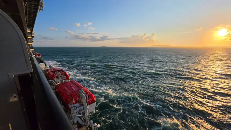 A view from the balcony of a Virgin Voyages cruise ship cabin showing the ocean and a sunset on the horizon. Lifeboats are visible along the side of the ship.