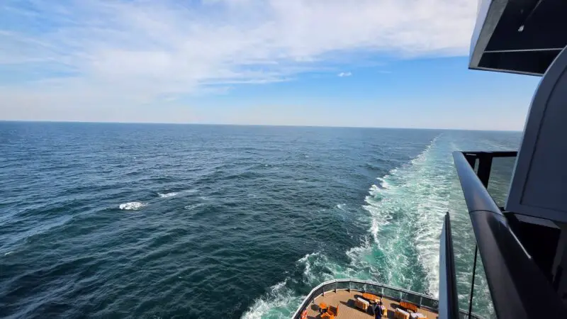 View from a Virgin Voyages cruise ship balcony overlooking the ocean, with lounge chairs and wake visible.