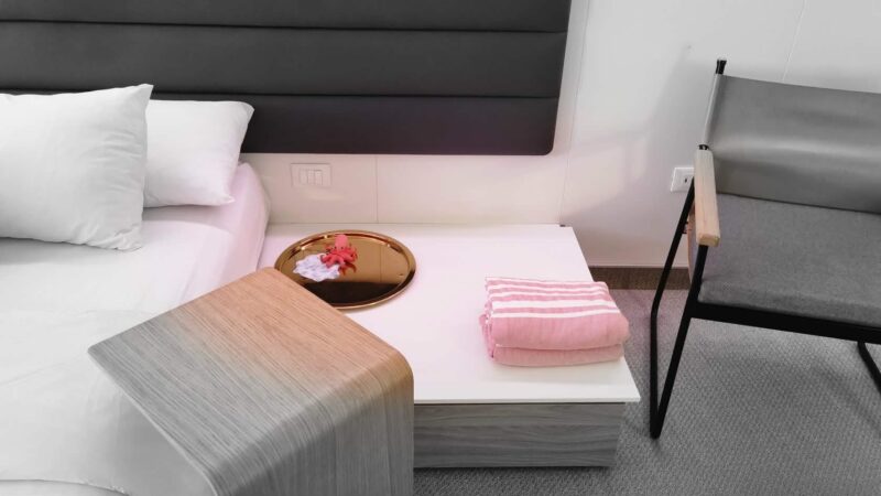 Virgin Voyages cruise ship cabin featuring a bed with white pillows, adjacent to a table with a pink folded towel and a decorative tray with a small red octopus figure. A gray chair is placed nearby.
