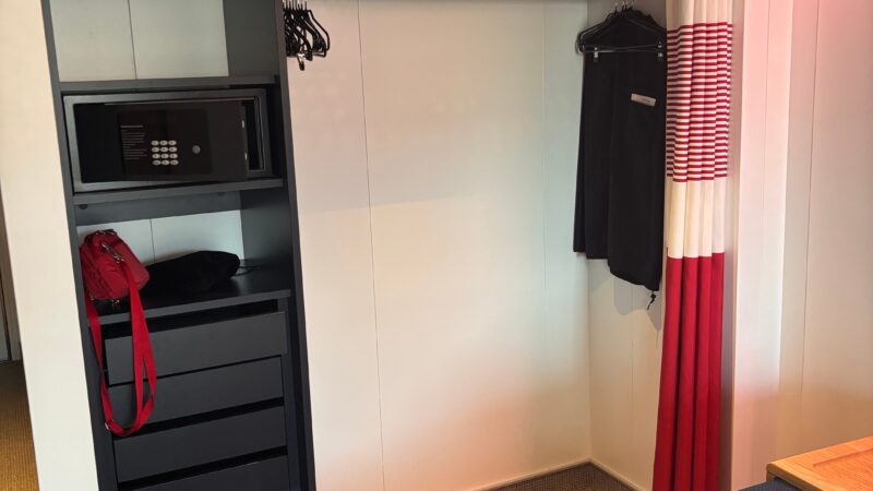 Virgin Voyages cruise ship cabin featuring a wardrobe with hangers, life jackets, a safe, and a curtain with red accents.