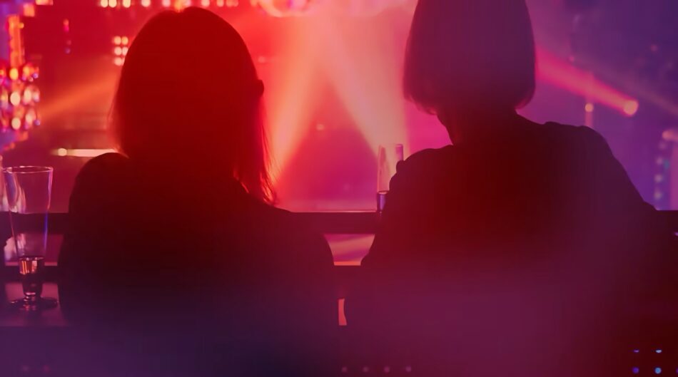 Two silhouetted people sit at a bar in a dim club with pink purple lighting