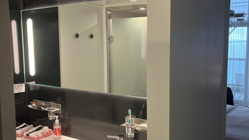 Interior of a Virgin Voyages cruise ship cabin bathroom with a modern sink, toiletries, and a lit mirror; a portion of the stateroom is visible in the background.