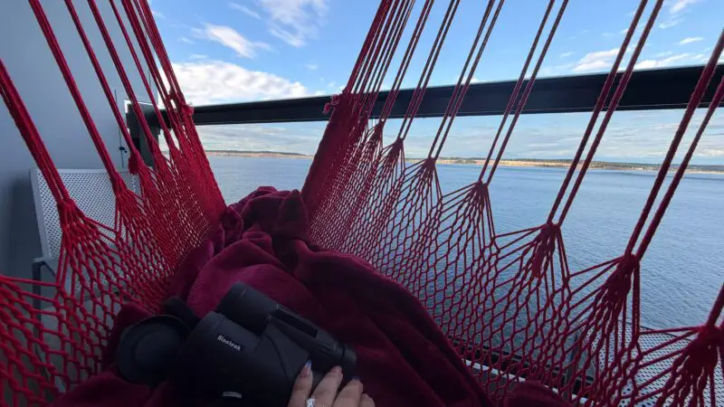 A cruise ship balcony with red hammock, blanket, binoculars, and ocean view.