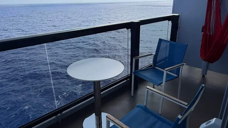 Balcony of a Virgin Voyages cruise ship cabin featuring two blue chairs, a small round table, and a red hammock, with an ocean view and blue sky in the background.