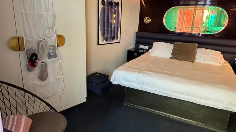 Virgin Voyages cruise ship cabin with a bed, porthole window, chair, and wall art.