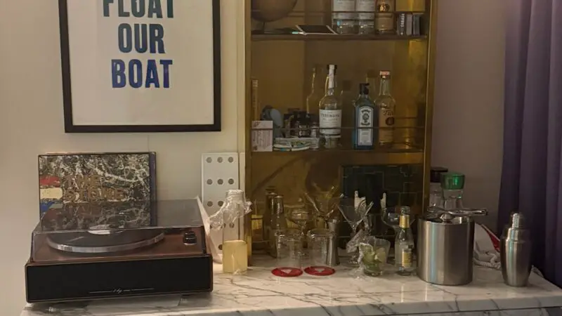 Virgin Voyages cruise ship cabin featuring a bar area with a marble countertop, turntable, bottles, and glasses, with a framed "You Float Our Boat" poster above.