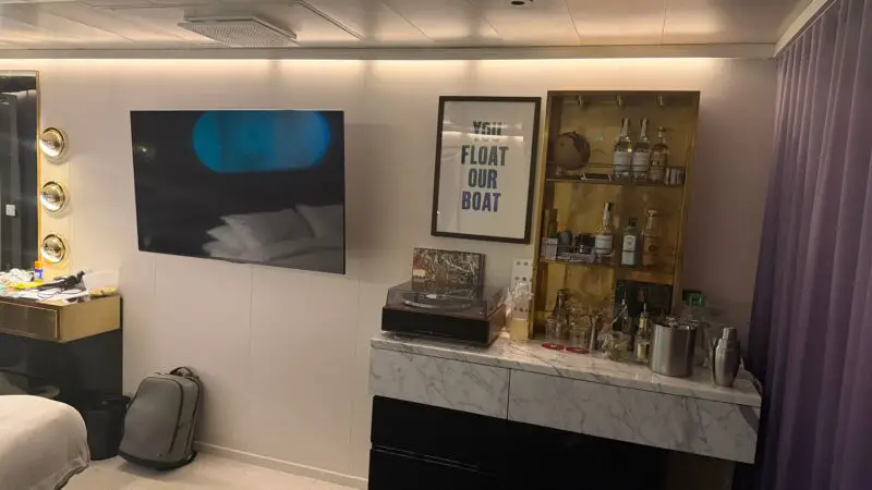 Virgin Voyages cruise ship cabin with a wall-mounted TV, bar area with various bottles, a framed sign, turntable, and purple curtains.