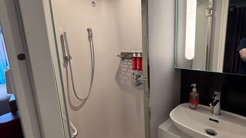Cruise ship cabin bathroom featuring a shower with a rainfall showerhead, sink with a lit mirror, and toiletries.